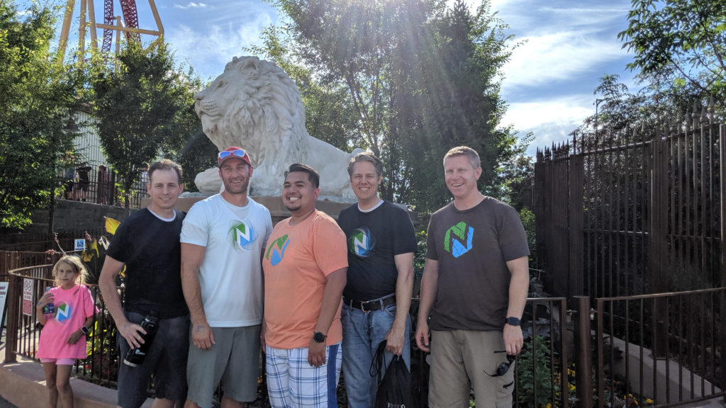 5 men wearing company tshirts standing in front of a metal fence. 
