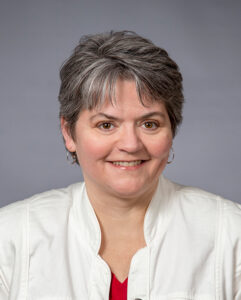 Caucasian woman with short salt and pepper hair, wearing a white jacket and red shirt.