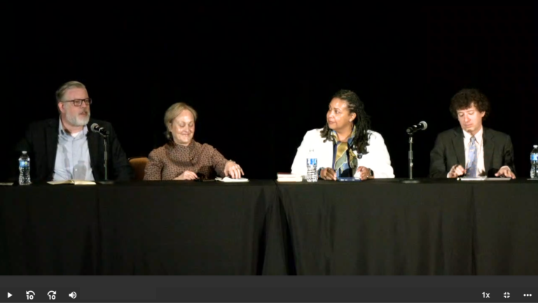 4 individuals sitting on a stage with black background and black draping. Individual 1, John Chraska, weating a black suit jacket and blue shirt, Individual 2 Kathleen McEvoy wearing a tan shirt, Individual 3 Nancy Kirkpatrick wearing a white suit jacket and multicolored shirt, Individual 4 Roger Schonfeld wearing a dark suit coat, light colored shirt, and blue necktie.