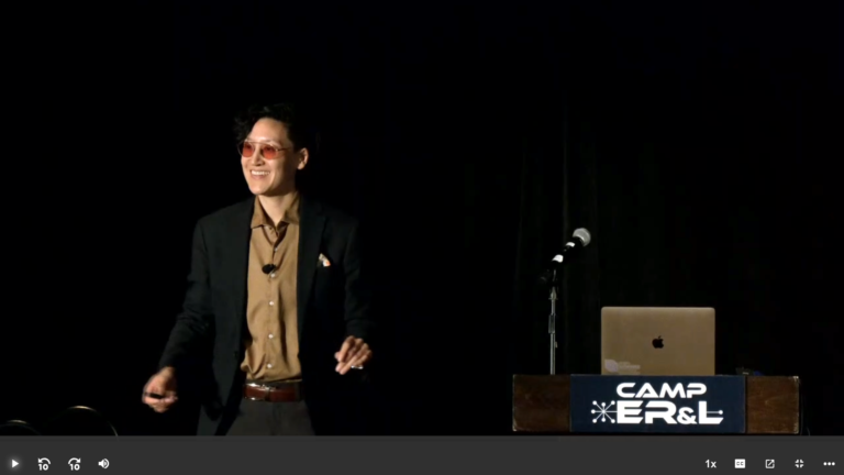 Kasia Chmielinski, a nonbinary person, wearing a black suit jacket, tan shirt, dark pants, and red tinted glasses standing next to a wooden podium with a laptop and microphone and Camp ER&L sign attached to the front.