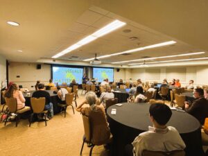 Attendees sitting around tables with black tablecloths, a presenter at the front of the room with 2 projection screens.